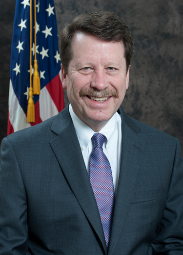 A headshot photo of Robert Califf. He is wearing a dark grey suit with a purple tie and white button up shirt. He is a white man with brown hair. He is smiling directly at the camera. The background has an American flag.